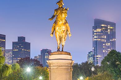 equestrian statue on a pedestal, set against a twilight cityscape with tall buildings and trees. the statue depicts a historical figure on horseback, illuminated against the dusky sky.