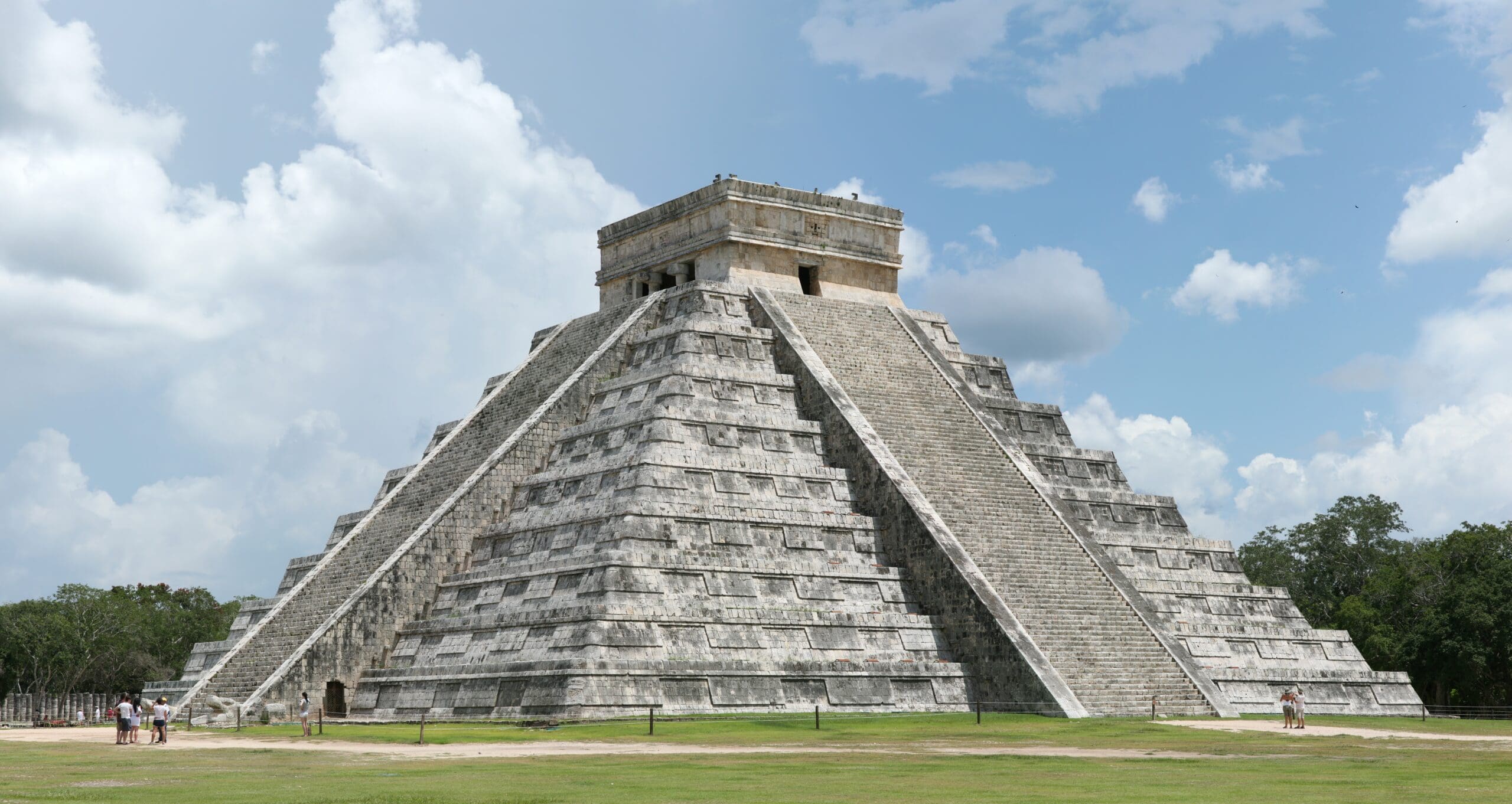 a large stone pyramid with a staircase on each side rises against a blue sky dotted with clouds. it stands on a grassy area, surrounded by scattered visitors. trees are visible in the background. a large stone pyramid with a staircase on each side rises against a blue sky dotted with clouds. it stands on a grassy area, surrounded by scattered visitors. trees are visible in the background.