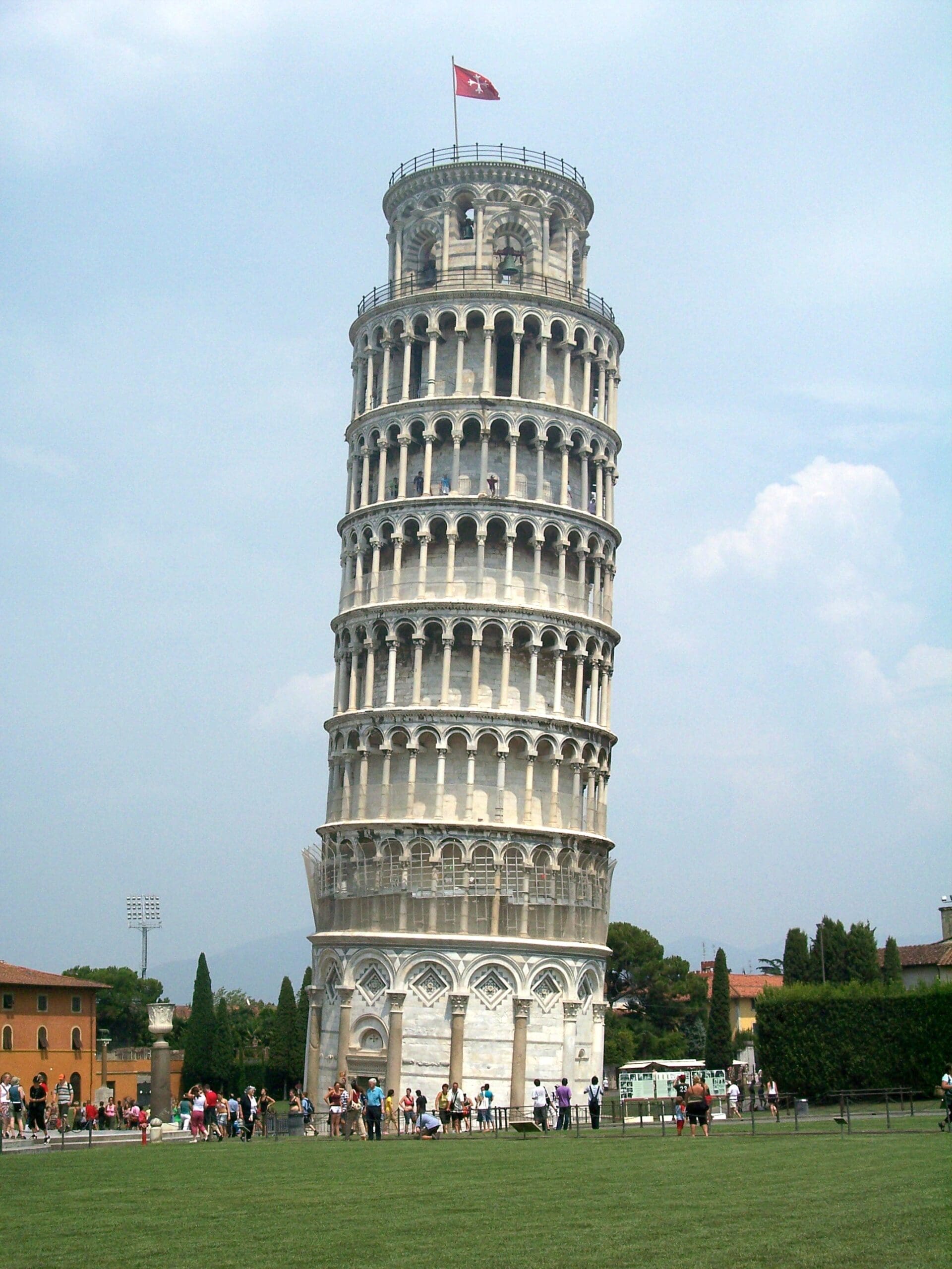 the image shows the leaning tower of pisa, a tall, cylindrical white marble bell tower with a noticeable tilt. it stands against a cloudy sky, surrounded by a lawn and tourists. a red flag flies atop the tower.